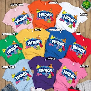 May include: A collection of colorful t-shirts in various shades, including light pink, orange, white, green, yellow, blue, red, and purple. Each shirt features the word "Nerds" in a playful font, accompanied by cartoon characters.