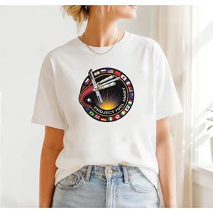 Puede incluir: Camiseta blanca con un diseño circular que dice "PROJECT HAIL MARY". El diseño incluye un cohete, un arco rojo, un sol y banderas de varios países. La camiseta es de cuello redondo.