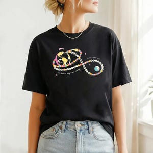 Puede incluir: Camiseta negra con un diseño gráfico colorido que presenta la Tierra, estrellas y los textos "It's been a long time coming" y "In our moon era". El diseño es de estilo caprichoso.