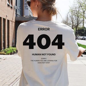 May include: Ivory-colored t-shirt with the text "ERROR 404 HUMAN NOT FOUND" in black. The shirt is worn by a person riding a scooter on a sidewalk. The text "SORRY THE HUMAN YOU ARE LOOKING FOR DOES NOT EXIST" is also printed on the shirt.