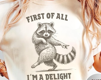 May include: An ivory t-shirt featuring a dark brown graphic illustration of a raccoon standing on its hind legs. The text above the raccoon reads FIRST OF ALL and the text below the raccoon reads I'M A DELIGHT in a bold, sans-serif font.
