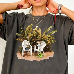 May include: Dark gray t-shirt featuring a tropical scene with two white chairs, banana leaves, and the text "Bad Bunny." The graphic tee has a relaxed fit and short sleeves. The shirt is a Pepper color.