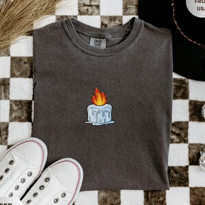 May include: A dark gray t-shirt with a small embroidered candle design. The candle has a blue base with a yellow and orange flame. The shirt is laid flat on a checkered surface, with white sneakers and a black hat in the background.