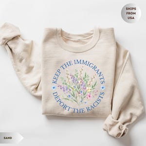 May include: Sand-colored sweatshirt with a floral design in the center. The design includes a circular text that reads "KEEP THE IMMIGRANTS, DEPORT THE RACISTS". The floral design features purple, blue, and pink flowers.