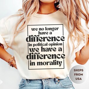 May include: Ivory-colored t-shirt with a black rectangular graphic containing the text: "we no longer have a difference in political opinion we have a difference in morality." The shirt is paired with blue jeans.