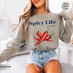 Op de afbeelding: Zandkleurig shirt met lange mouwen met de tekst "Spicy Life Everyday" boven een afbeelding van drie rode chilipepers. Het shirt heeft een neutrale kleur en een relaxte pasvorm.
