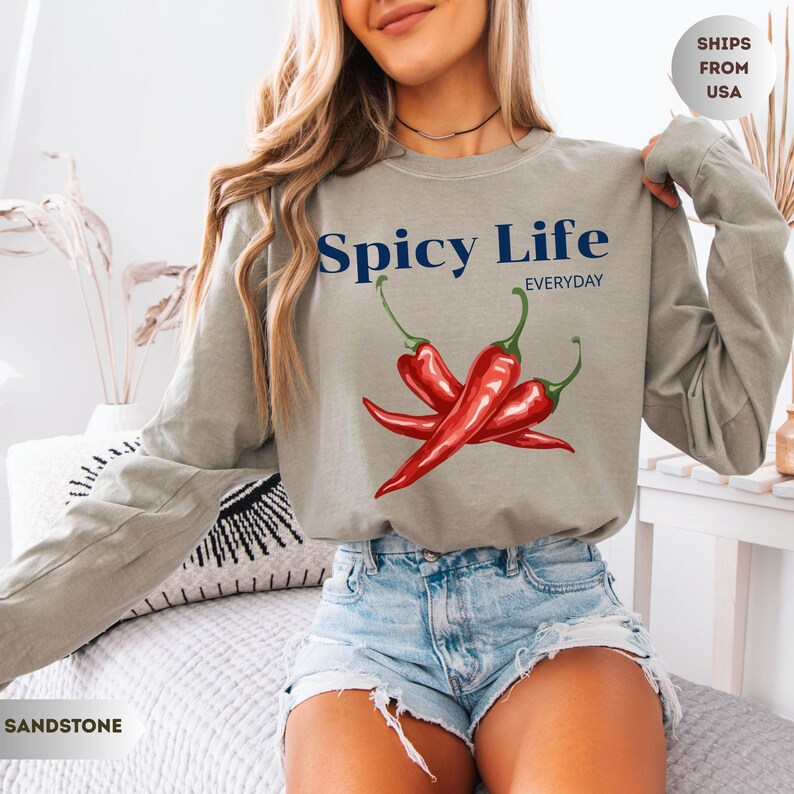Op de afbeelding: Zandkleurig shirt met lange mouwen met de tekst "Spicy Life Everyday" boven een afbeelding van drie rode chilipepers. Het shirt heeft een neutrale kleur en een relaxte pasvorm.