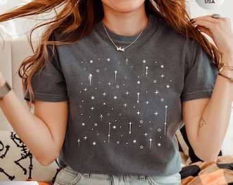 May include: A charcoal gray short-sleeve t-shirt featuring a white graphic print of various stars and shooting stars scattered across the chest. The shirt has a crew neckline and a relaxed fit, showcasing a celestial pattern of simple line-drawn stars.