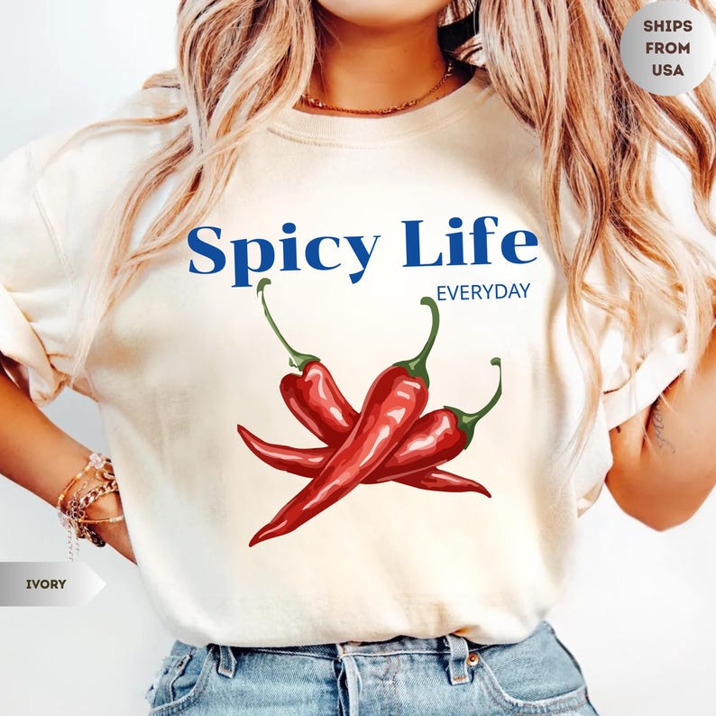 Op de afbeelding: Een ivoorkleurig T-shirt met korte mouwen en een afbeelding van drie rode chilipepers met groene steeltjes. De tekst "Spicy Life" staat in het blauw boven de pepers, met het woord "EVERYDAY" in kleinere blauwe letters eronder.