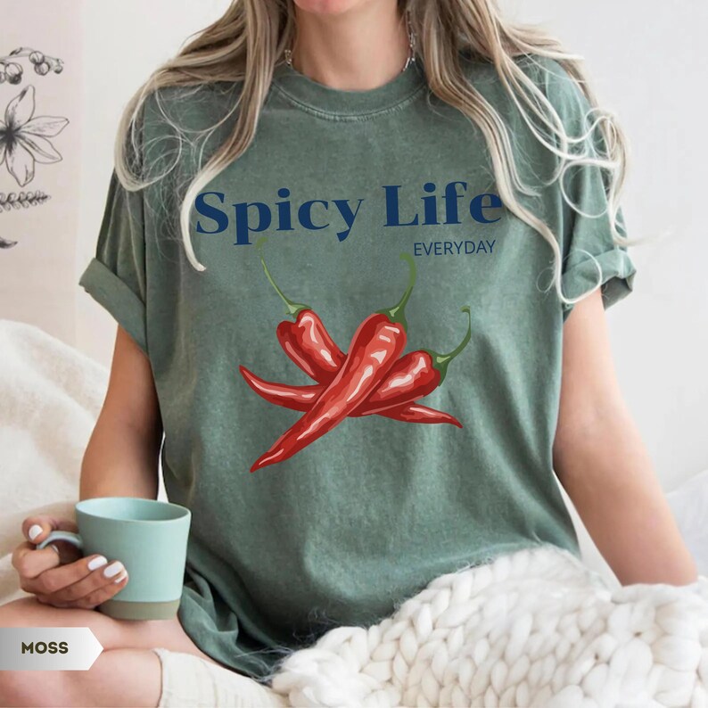 Op de afbeelding: Een mosgroen T-shirt met korte mouwen en de donkerblauwe tekst Spicy Life EVERYDAY boven een grafische illustratie van drie felrode chilipepers met groene steeltjes. Het T-shirt heeft een ronde hals en een relaxte pasvorm.