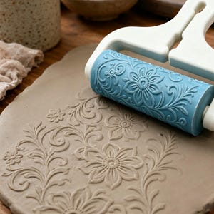May include: A light blue patterned clay roller with a white handle, shown pressing a detailed floral and vine design into a flat piece of gray clay. The roller features a repeating embossed motif of flowers and swirling leaves.