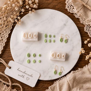 May include: Two white rectangular clay cutters with the text BORULA, featuring leaf and teardrop shapes. Includes several small light green clay leaf cutouts and two pairs of gold-toned hook earrings with green leaf charms and small white beads.