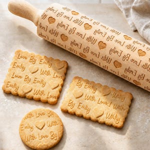 May include: A light-colored wooden rolling pin with an engraved pattern of hearts and the text "With Love By Emily" repeated across its surface, accompanied by three baked cookies featuring the same embossed heart and text design.