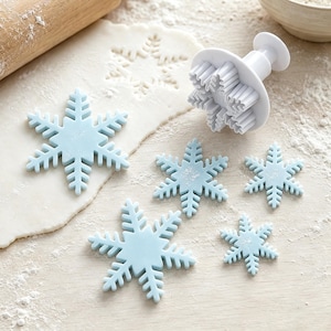 May include: A white plastic snowflake plunger cutter and several light blue fondant snowflake cutouts of varying sizes. The plunger tool features a spring-loaded mechanism with a detailed snowflake-shaped edge for pressing and cutting shapes.
