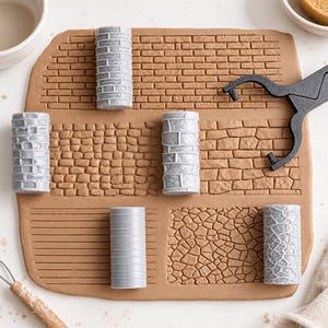 Op de afbeelding: Een set van zes cilindrische grijze plastic textuurrollers voor klei, elk met een uniek reliëfpatroon zoals baksteen, steen en horizontale lijnen, gedrukt in een platte plaat van lichtbruine boetseerklei met een zwart gereedschap.