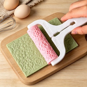 May include: A pink patterned rolling pin with a white handle featuring the text "This side up" embossed on the frame. The roller is pressing a floral design into a rectangular slab of light green dough, creating a detailed embossed texture.