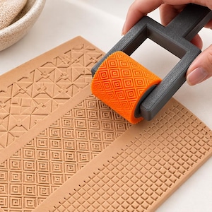Può includere: Un rullo in plastica grigio scuro con un cilindro arancione testurizzato caratterizzato da un motivo a quadrati ripetuti. Il rullo viene premuto su una superficie piana color cuoio, creando un design in rilievo di piccoli quadrati.