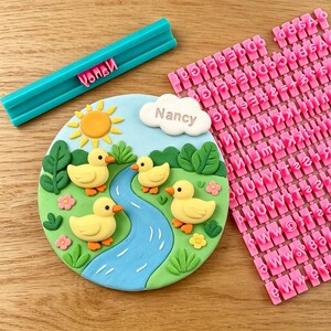 May include: A colorful cake decoration featuring four yellow ducklings near a blue river, with a sun and cloud that says "Nancy". Includes a teal letter press and pink letter stamps for customization.