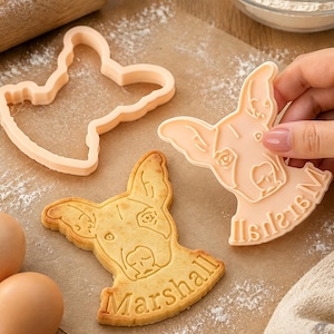May include: A light peach-colored plastic cookie cutter shaped like a dog head with the name Marshall embossed at the bottom, alongside a baked cookie featuring the same dog face and the name Marshall imprinted in the dough surface.