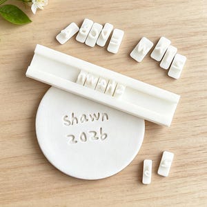 May include: A white plastic letter stamp set featuring individual character tiles and a long rectangular holder. A circular white clay disc is imprinted with the text Shawn 2026, created using the stamp set. The letters are in a sans-serif font.