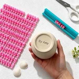 Pode incluir: Um conjunto de carimbos de alfabeto rosa, um suporte de carimbo turquesa e uma pequena tigela de cerâmica. A tigela tem o texto "By Amy" impresso. Duas conchas e uma tesoura também estão na imagem.