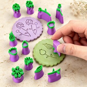 May include: A collection of purple and green flower-shaped cookie cutters and stamps. The set includes various floral designs, such as roses, tulips, and leaves. A hand is pressing a cutter onto a green cookie.