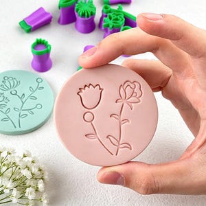 May include: A hand holds a pink, circular mold with embossed floral designs, including a tulip and a rose. Other purple and green molds are in the background. A light green mold with a floral design is also visible.
