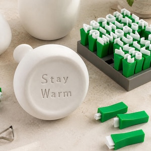 May include: A white circular ceramic stamp featuring the embossed text "Stay Warm" on its face, accompanied by a grey tray containing a set of green and white plastic alphabet letter stamps and three loose green and white letter stamps.