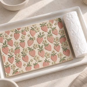 Puede incluir: Una placa de arcilla rectangular con un patrón en relieve repetido de fresas rosas con hojas verdes y flores verde claro, acompañada de un rodillo cilíndrico blanco a juego con el mismo motivo de fresas y flores.