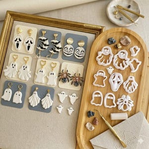 May include: A collection of Halloween-themed earrings and crafting tools. The earrings feature ghost, bat, spider, and jack-o'-lantern designs. The tools include cutters, a stylus, and clay. The earrings are displayed on gray and beige cards.