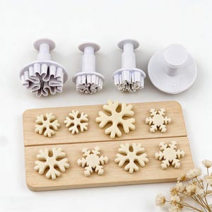 May include: A set of white plastic snowflake cookie cutters and several snowflake-shaped cookies on a wooden board. The cutters have a plunger design for easy use. The cookies are light brown.