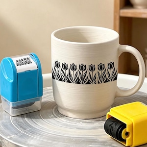 May include: A cream-colored ceramic mug featuring a horizontal band of black tulip illustrations. Beside the mug are two plastic rolling stamps, one bright blue and one bright yellow, used for applying the black tulip pattern to the surface.