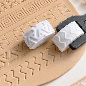 Op de afbeelding: Twee witte cilindrische kleirollers met verhoogde geometrische patronen, waarvan er één aan een zwarte kunststof handgreep is bevestigd, rustend op een beige kleiplaat met ingedrukte Griekse, visgraat- en zigzagontwerpen.