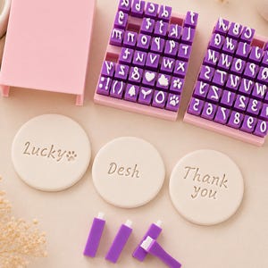 以下が含まれることがあります： 2つのピンクのトレイと蓋に収められた、文字と数字の紫色のゴム印セット。Lucky、Desh、Thank youという言葉が刻印された3つの白い円形の粘土ディスクと、いくつかの紫色のスタンプハンドルが配置されています。
