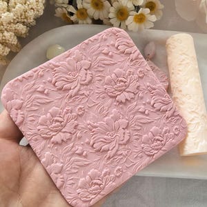 May include: A square, pink, floral-patterned object with a detailed embossed design of peonies and leaves. A cylindrical object with a similar floral pattern is next to it. The objects are on a white surface with flowers in the background.