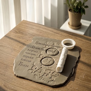 May include: A gray clay slab with embossed text and two circular impressions. A white rolling pin and a circular cutter sit on the slab. The text is in a script font. The items are on a wooden table.