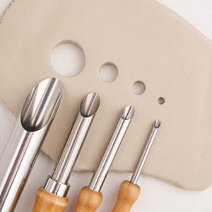May include: A set of four stainless steel clay hole cutters with wooden handles, arranged in descending order of size. Each tool features a sharp, cylindrical metal tip designed for punching circular holes into a flat piece of light beige clay.