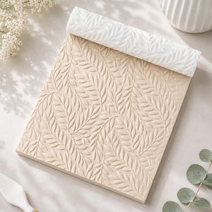 May include: A rectangular beige clay slab featuring a debossed leaf pattern across its surface, paired with a white cylindrical rolling pin that displays a matching raised leaf design, both used for creating textured impressions in crafting materials.