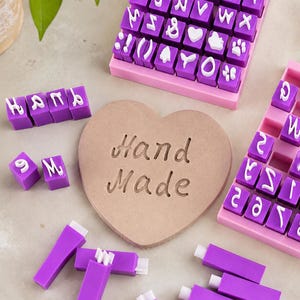 Puede incluir: Un conjunto de sellos de letras y símbolos en color morado y blanco organizados en bandejas rosas, junto a sellos individuales y una pieza de arcilla beige con forma de corazón que muestra el texto Hand Made en letra cursiva.