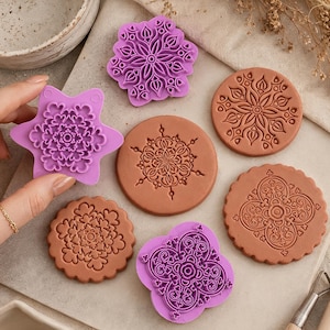May include: A collection of purple plastic stamps and terracotta-colored clay discs featuring intricate, symmetrical geometric and floral patterns. The stamps have raised designs for pressing into clay, while the clay discs show the embossed impressions.