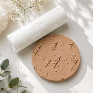 May include: A white cylindrical rolling pin with a debossed pattern of pine trees and small dots, positioned next to a circular tan clay disc that features the same embossed pine tree and dot pattern pressed into its flat surface.