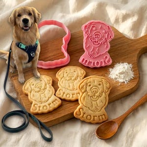 Op de afbeelding: Een Golden Retriever-hond zit naast hondenkoekjes en een roze koekjesvorm. De koekjes zijn gestempeld met de naam "Storm". Een houten lepel en een hoop bloem liggen ook op de snijplank.