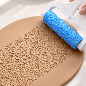 May include: A blue patterned rolling pin with a white handle featuring the text "This side up" embossed on the frame. The roller is pressing a repeating floral and swirl design into a flat, tan-colored piece of clay, creating a textured surface.