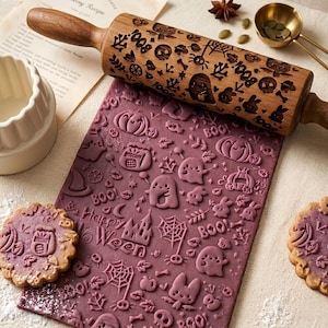 May include: A wooden rolling pin with engraved Halloween motifs including ghosts, pumpkins, spiders, and the text 'BOO'. The rolling pin is positioned above a sheet of purple dough embossed with matching patterns, alongside two round cookies.