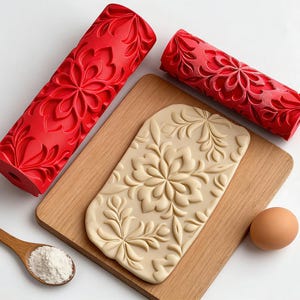 Peut inclure: Deux rouleaux à pâtisserie rouges à motifs floraux et une pâte avec un motif floral en relief sur une planche en bois. Une cuillère en bois avec de la farine et un œuf sont également présents.
