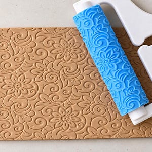 Può includere: Un mattarello testurizzato blu brillante con telaio a manico bianco, posizionato sopra un foglio rettangolare color marrone chiaro che presenta un motivo floreale e a volute in rilievo che corrisponde al design del rullo.