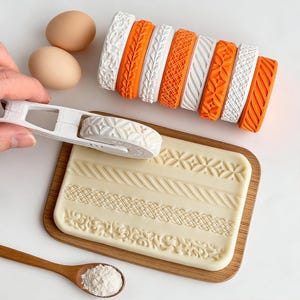 May include: A white rolling pin with a patterned roller is held over a rectangular mold with various embossed designs. Several orange and white patterned rollers are displayed, along with two eggs and a wooden spoon with flour.