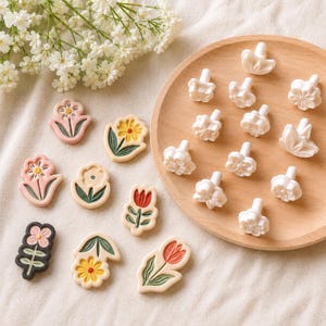 May include: A collection of flat, colorful flower-shaped clay pieces and white, three-dimensional flower-shaped stamps. The flat pieces feature various floral designs in pink, yellow, red, and green. The white stamps have handles.