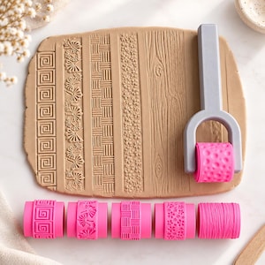Op de afbeelding: Een lichtgrijze kunststof handgreep met een afneembare roze rolkop, vergezeld van vijf verwisselbare roze patroonrollen. De rollen creëren texturen op een plat stuk beige klei, waaronder geometrische, florale en houtnerfpatronen.
