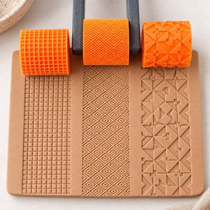 Puede incluir: Un juego de tres rodillos cilíndricos de color naranja con distintos patrones geométricos, incluyendo una cuadrícula, diamantes y triángulos, montados sobre un mango gris oscuro. Debajo hay una placa rectangular de color canela.